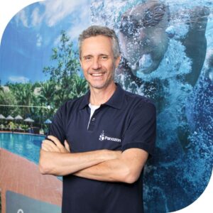 É uma foto em plano médio, com as bordas arredondadas. O assunto principal é um homem adulto, identificado pelo nome do arquivo como Carlos. A paisagem é um banner ou backdrop com uma imagem de piscina e uma pessoa submersa, mergulhando. A ação é o homem está em pé, sorrindo para a câmera com os braços cruzados. Ele usa uma camisa polo azul-marinho com a logomarca da "Panozon" bordada no lado esquerdo do peito. Fim de descrição.