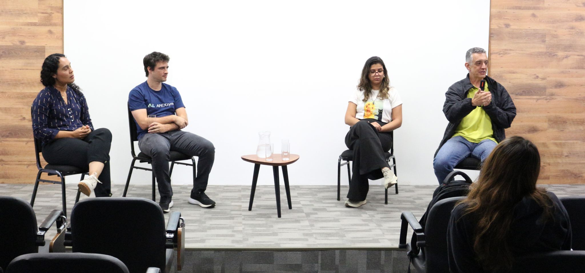 Quatro pessoas participam de uma roda de conversa em um auditório. Elas estão sentadas em cadeiras dispostas em linha, em frente a uma parede branca com detalhes em madeira. A ordem é, de uma extremidade a outra: uma mulher, um homem, uma mulher e um homem. No meio do palco, há uma pequena mesa redonda com copos e uma jarra de água. Na frente deles, algumas pessoas do público acompanham a atividade. Fim da descrição.