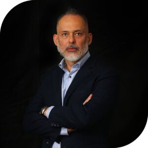 É uma foto em plano médio, com as bordas arredondadas. O assunto principal é um homem adulto, identificado pelo nome do arquivo como Iron. A paisagem é um fundo preto liso, com iluminação de estúdio que destaca o rosto e o tronco. A ação é o homem está posando para a câmera com os braços cruzados. Ele usa um blazer escuro sobre uma camisa social clara. Ele tem barba e bigode grisalhos e olha para a câmera com uma expressão séria. Fim de descrição.