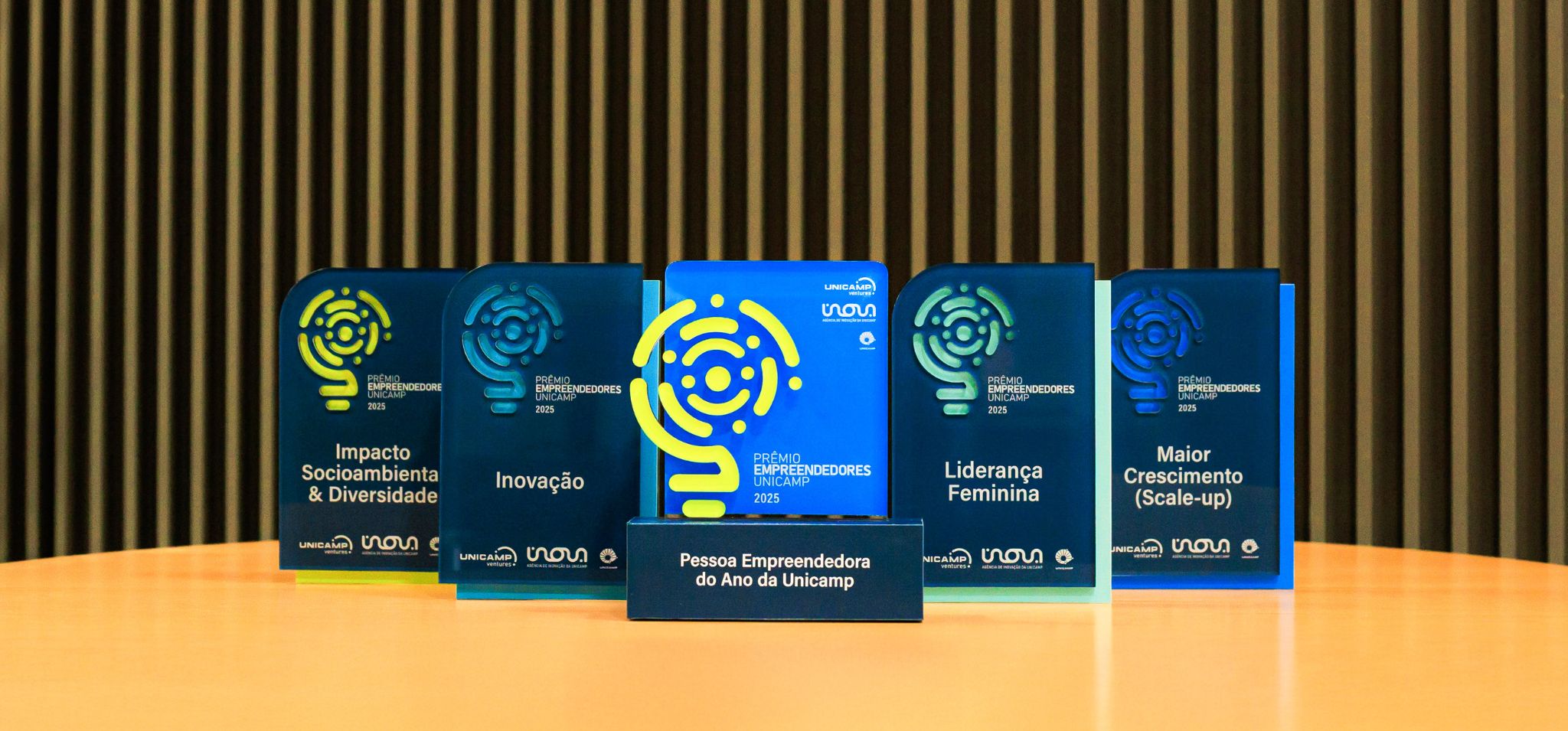 É uma foto em plano detalhe dos troféus de um prêmio. O assunto principal são cinco troféus do Prêmio Empreendedores Unicamp 2025. A paisagem é um ambiente interno com um fundo de painel de madeira com listras verticais. Os troféus estão dispostos sobre uma mesa ou balcão de madeira. A ação é de exibir os troféus, que são feitos de acrílico azul-escuro com detalhes em amarelo-limão e azul claro. Cada um está rotulado com uma categoria: Impacto Socioambiental & Diversidade, Inovação, Liderança Feminina e Maior Crescimento (Scale-up). O troféu central, maior e azul-claro, representa a categoria "Pessoa Empreendedora do Ano da Unicamp". Fim de descrição.