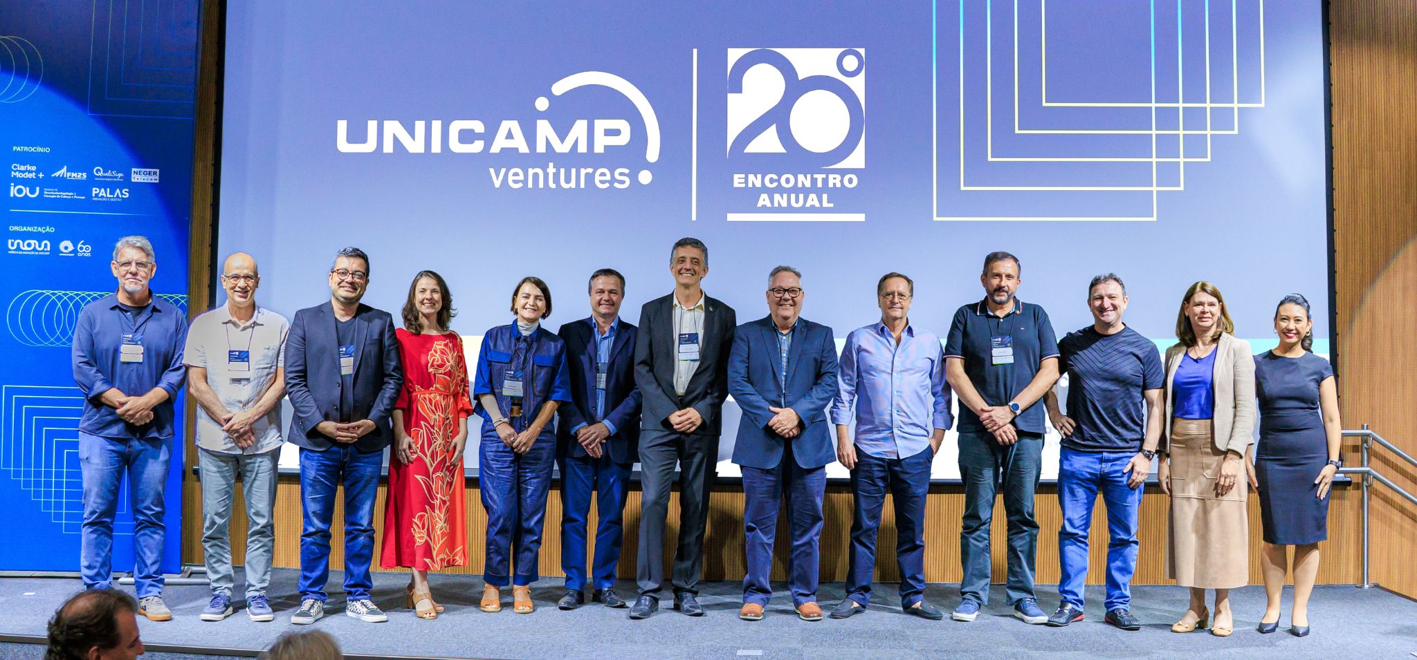 Fotografia em ambiente de auditório, mostrando um grupo de pessoas alinhadas no palco durante o 20º Encontro Anual Unicamp Ventures, diante de um telão com a identidade visual do evento. Fim da descrição.