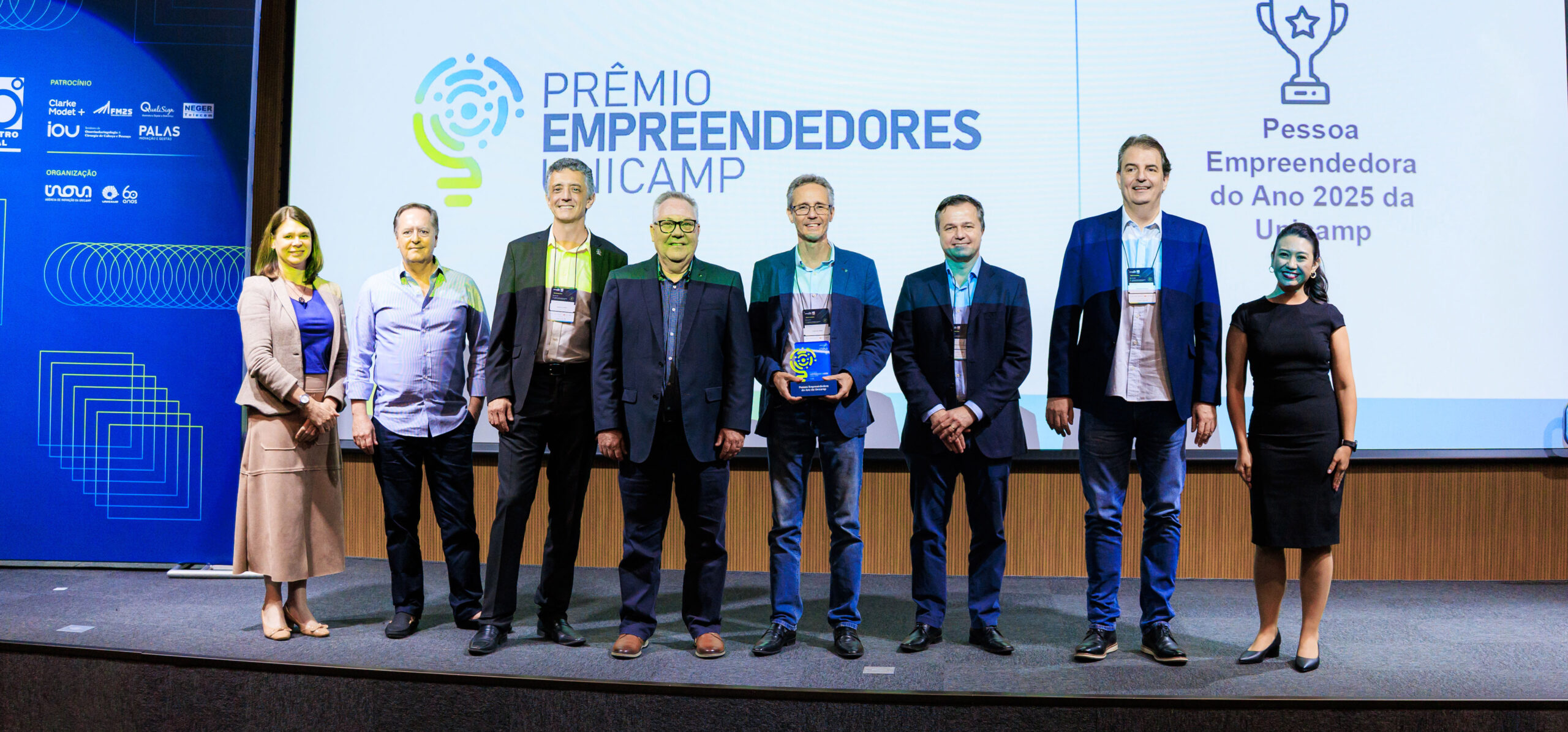 Fotografia em ambiente de auditório, mostrando um grupo de oito pessoas alinhadas no palco durante a cerimônia do Prêmio Empreendedores Unicamp, com um dos participantes segurando o troféu diante de um telão com a identidade visual do evento. Fim da descrição.