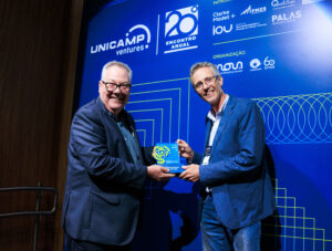Fotografia em ambiente de palco de evento, mostrando dois homens sorrindo enquanto um deles entrega um troféu do Prêmio Empreendedores Unicamp 2025 ao outro diante de um painel azul com a identidade visual do 20º Encontro Anual Unicamp Ventures. Fim da descrição.