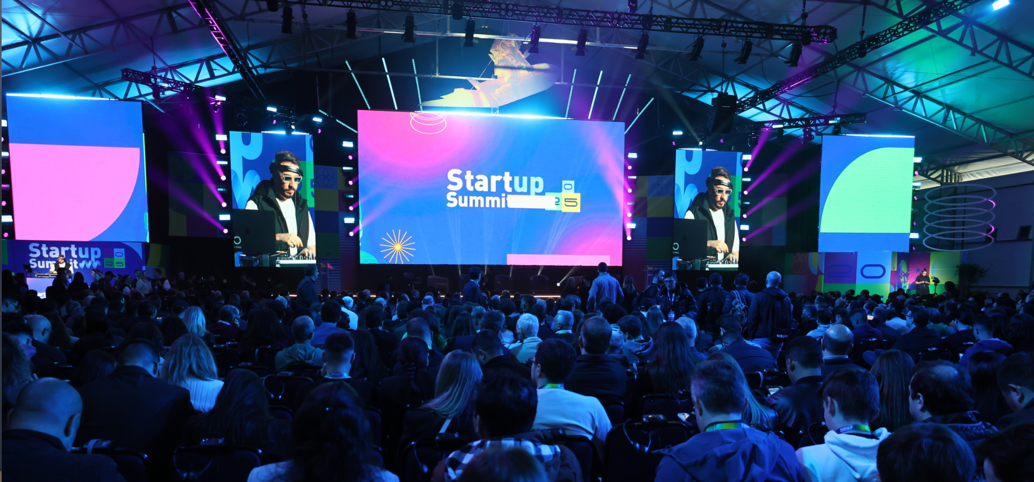 Foto colorida apresenta um espaço de eventos lotado, com luzes coloridas. Fim da descrição