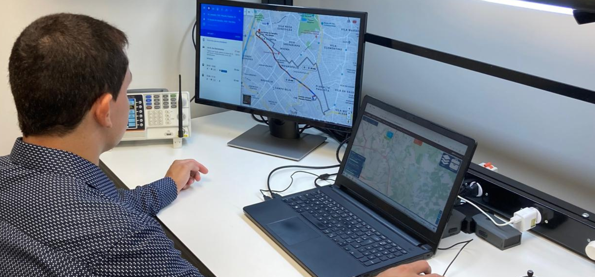 Foto colorida mostra um homem observando a tela de dois computadores. Fim da descrição.
