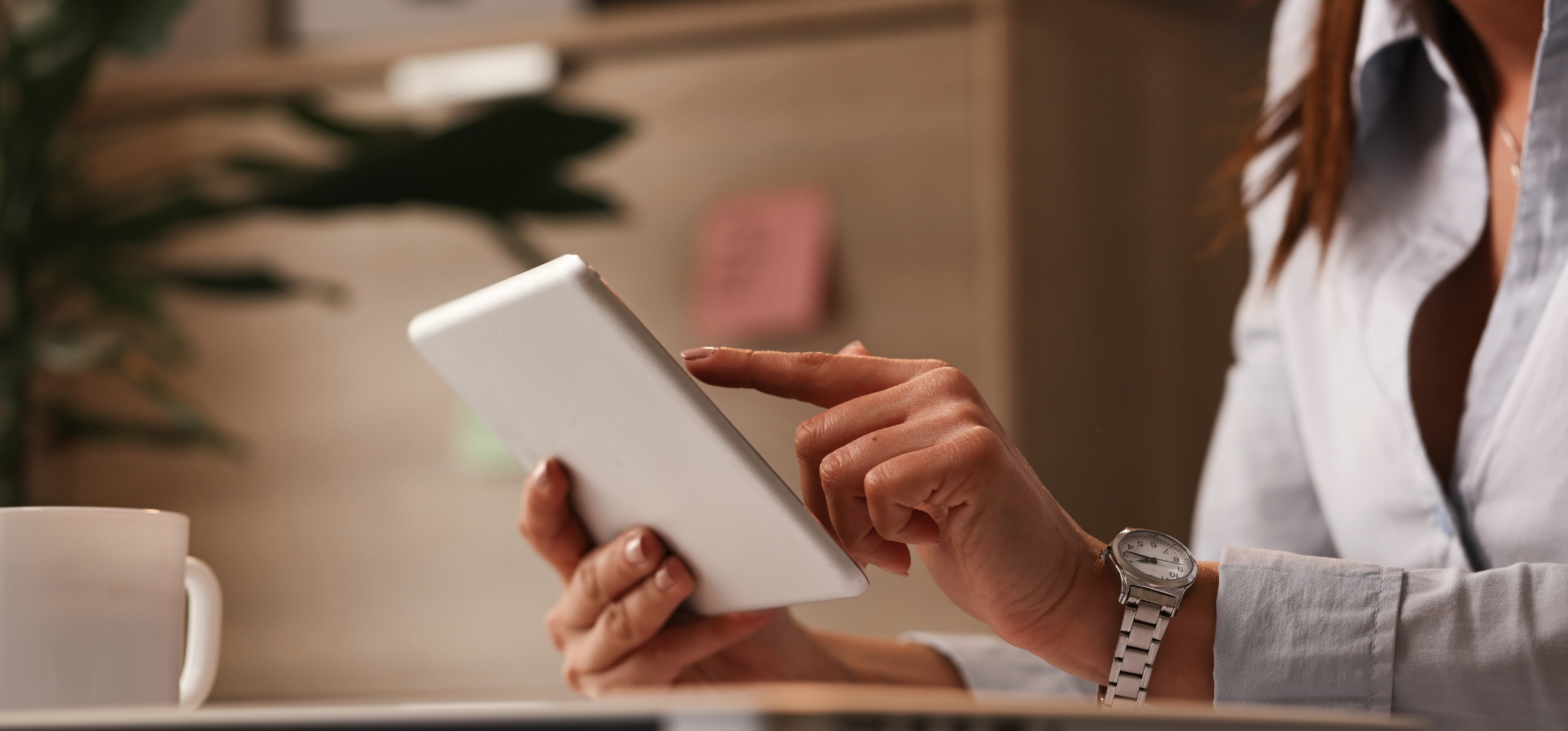 Imagem colorida apresenta uma mulher manuseando um dispositivo digital em um ambiente escuro e com plantas, similar a uma sala de estar. Fim da descrição.