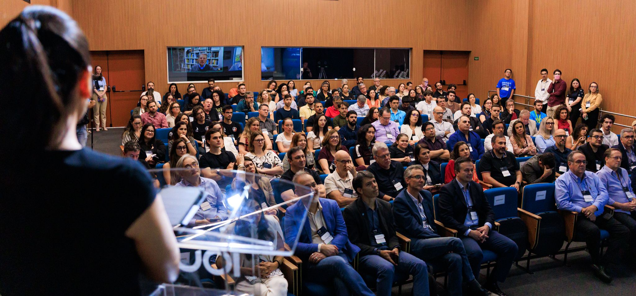 Aqui está a descrição da imagem, seguindo o formato e as restrições solicitadas (sem negritos, menos de 1000 caracteres): É uma foto de uma grande plateia em um auditório. A cena acontece em um ambiente interno (auditório) com fileiras de assentos azuis e paredes de madeira escura com painéis verticais. Há duas telas quadradas na parede do fundo que exibem uma vídeo-chamada com um palestrante. A iluminação é de evento. A ação é a plateia, composta por muitas pessoas adultas vestidas em trajes casuais de negócios, está sentada nas poltronas, observando o palco ou a pessoa que está discursando. Em primeiro plano, uma mulher de costas, usa uma camiseta preta e está em pé atrás de um púlpito transparente, de frente para o público. Selo de empresa-filha da Unicamp. Fim de descrição.
