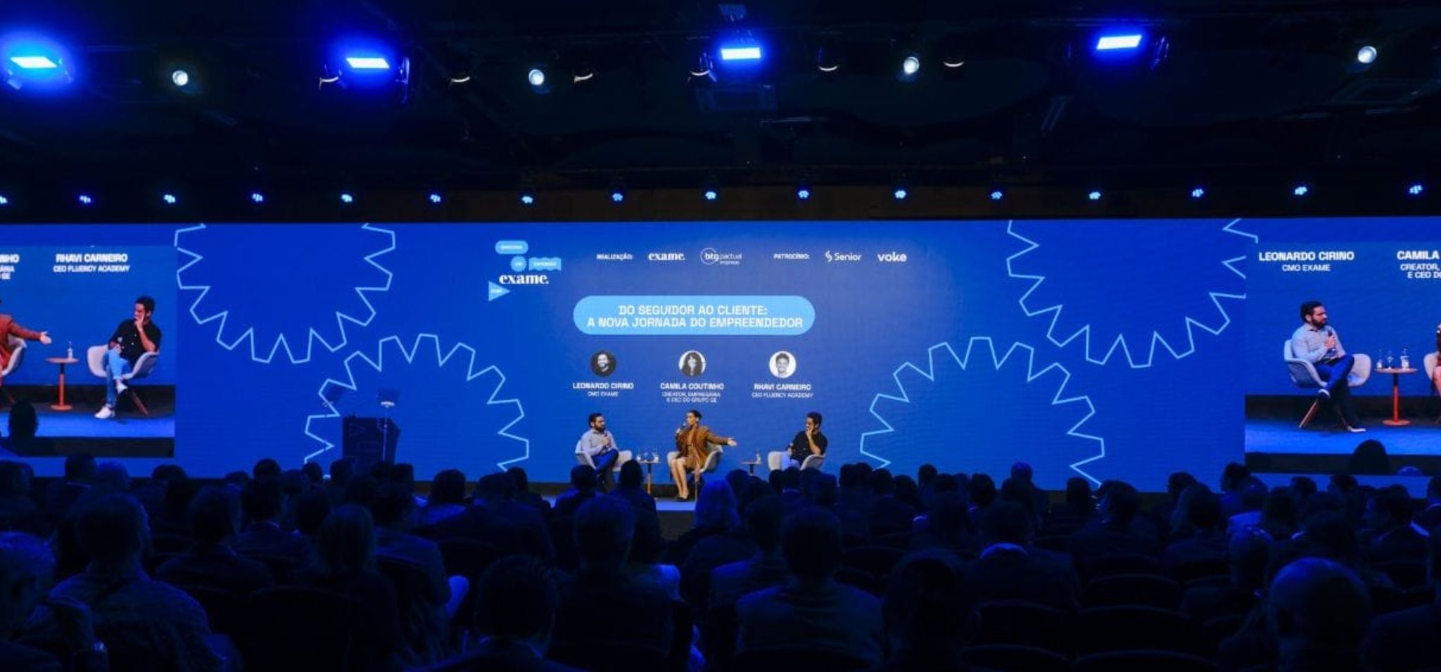 É uma foto horizontal de um palco de evento em um auditório. O assunto principal são três pessoas (dois homens e uma mulher) sentadas em poltronas participando de uma mesa redonda. A paisagem é o palco de um evento corporativo com um telão gigante de fundo na cor azul que exibe grafismos de engrenagens brancas e o título "DO SEGUIDOR AO CLIENTE: A NOVA JORNADA DO EMPREENDEDOR". No telão também aparecem as fotos e nomes dos palestrantes: Leonardo Cirino, Camila Coutinho e Rhavi Carneiro, além dos logotipos da Exame, BTG Pactual, Senior e Voke. A ação é que os palestrantes estão sentados em poltronas brancas no centro do palco, participando de uma discussão. Em primeiro plano, a plateia é vista em silhueta escura, de costas para a câmera, observando a apresentação. empresas-filhas Fim de descrição.