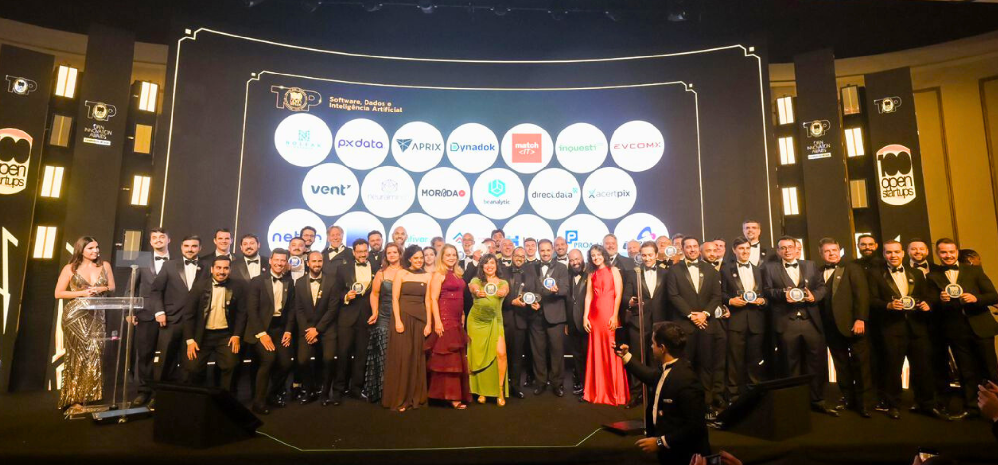 É uma foto de um grande grupo de pessoas em um evento de gala. O assunto principal é um grupo de cerca de 40 pessoas (homens e mulheres) em um palco, recebendo um prêmio. A paisagem é o interior de um salão de eventos elegante. O fundo é um grande painel de LED ou tela que exibe o logo "TOP 100 Open Startups" e lista vários logotipos de empresas sob o título "Software, Dados e Inteligência Artificial". O painel tem detalhes arquitetônicos em arco iluminado. 100 Open Startups. A ação é o grupo está em pé no palco, posando para a foto, a maioria segurando troféus ou prêmios. A maioria dos homens usa smoking preto com gravata borboleta, enquanto as mulheres usam vestidos de festa coloridos (tons de dourado, vermelho, verde e marrom). Fim de descrição.