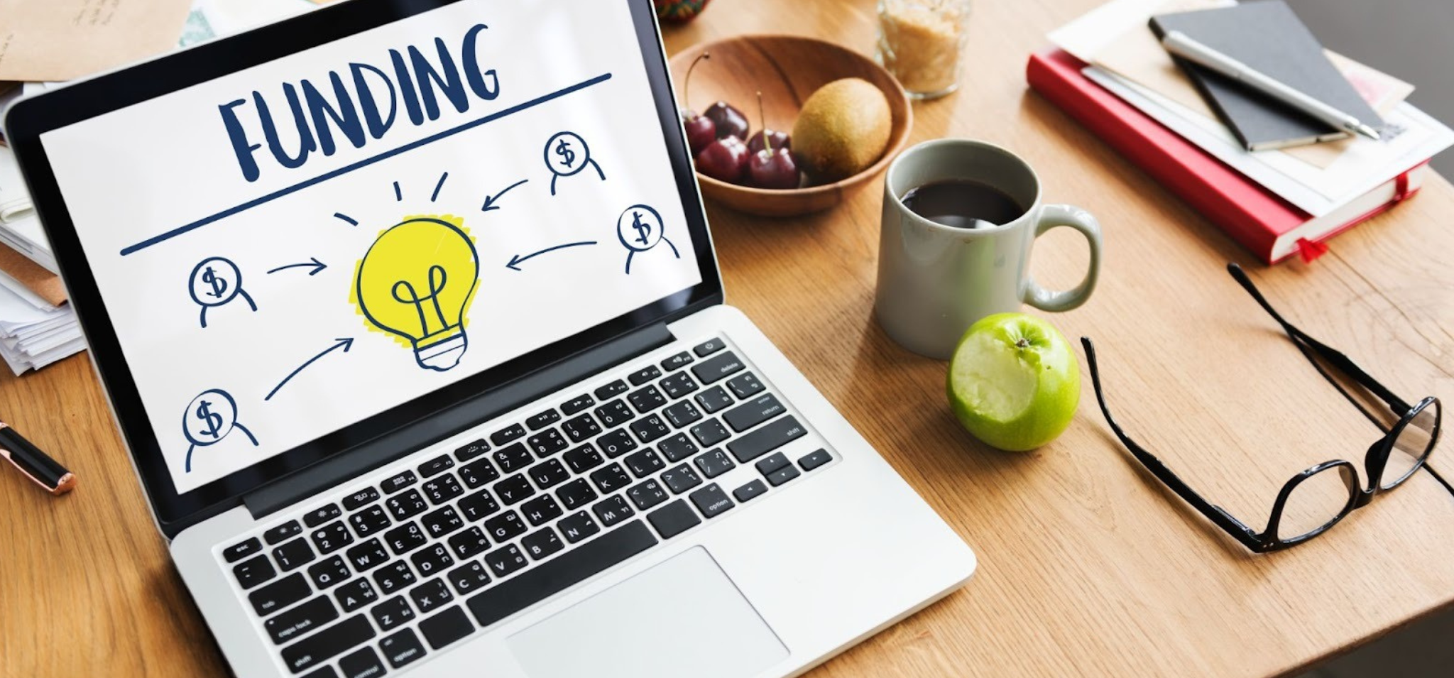 Foto colorida apresenta um notebook posicionado ao lado de algumas frutas e uma xícara de café. Fim da descrição.