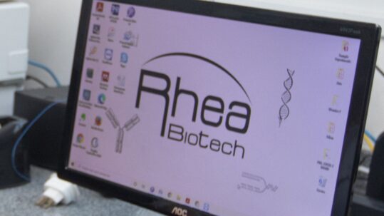 Foto de um monitor de computador exibindo o logotipo da Rheabiotech, acompanhado de ícones de uma hélice de DNA e de um anticorpo. O monitor está em um ambiente de laboratório com equipamentos ao fundo. Fim da descrição.