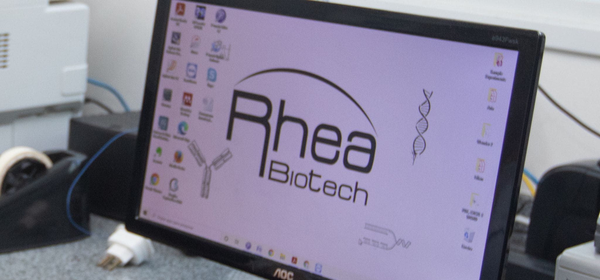Foto de um monitor de computador exibindo o logotipo da Rheabiotech, acompanhado de ícones de uma hélice de DNA e de um anticorpo. O monitor está em um ambiente de laboratório com equipamentos ao fundo. Fim da descrição.