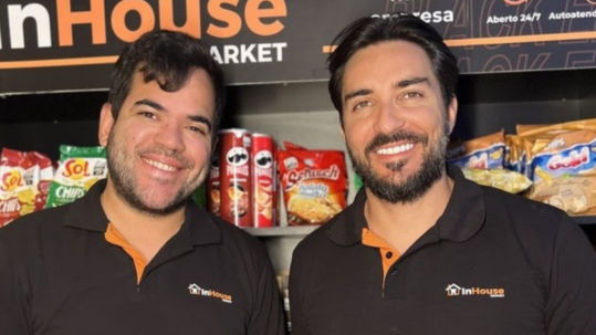 Foto colorida apresenta dois homens sorrindo para a foto, em frente a prateleiras de mercado. O homem à esquerda possui barba rala e o da direita, barba cheia e bem aparada. Ao fundo, veem-se diversas embalagens coloridas de salgadinhos e, na parte superior, um painel com o nome da empresa e as informações "Aberto 24/7" e "Autoatendimento". Fim da descrição.