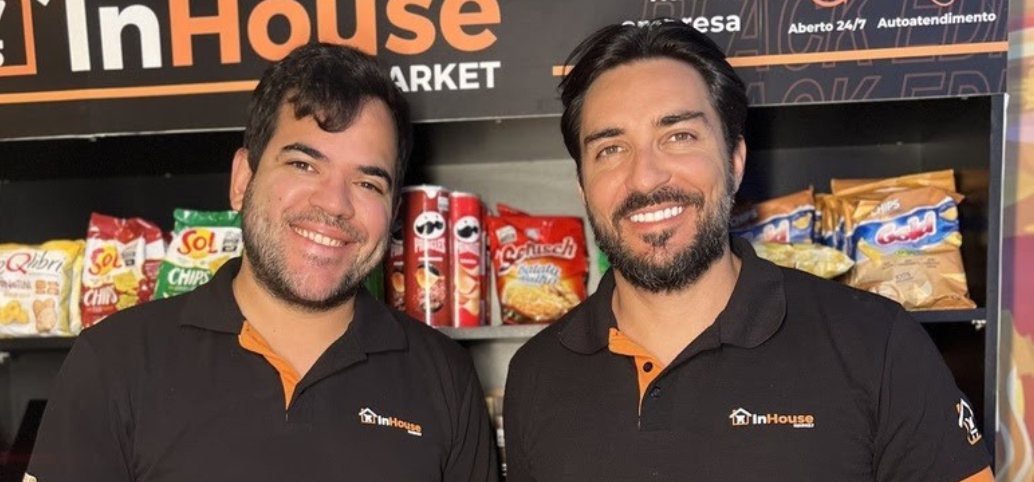 Foto colorida apresenta dois homens sorrindo para a foto, em frente a prateleiras de mercado. O homem à esquerda possui barba rala e o da direita, barba cheia e bem aparada. Ao fundo, veem-se diversas embalagens coloridas de salgadinhos e, na parte superior, um painel com o nome da empresa e as informações "Aberto 24/7" e "Autoatendimento". Fim da descrição.