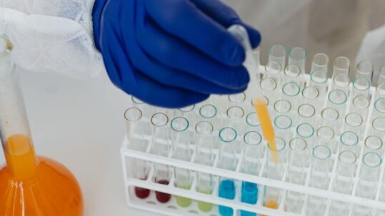 Foto colorida apresenta uma mão manuseando frascos de laboratórios contando substâncias líquidas coloridas. Fim da descrição.