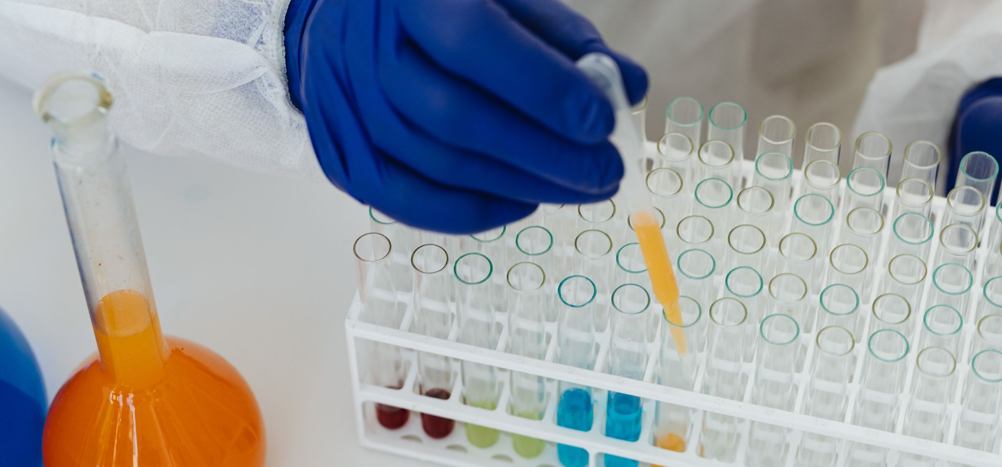 Foto colorida apresenta uma mão manuseando frascos de laboratórios contando substâncias líquidas coloridas. Fim da descrição.