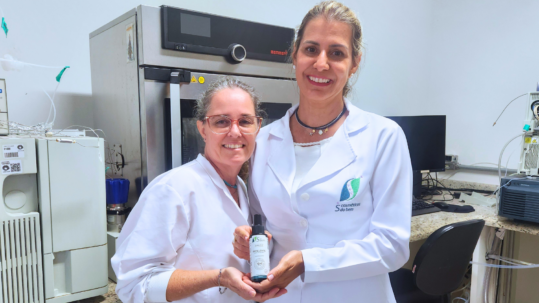 A imagem apresenta duas mulheres sorridentes, de peles claras, em um ambiente de laboratório, posando lado a lado e segurando juntas um frasco de produto cosmético. A mulher à esquerda tem cabelos grisalhos presos, usa óculos de armação transparente e veste um jaleco branco. A mulher à direita tem cabelos loiros longos, usa um colar escuro com pequenos pingentes e veste um jaleco branco com o logotipo "S do Bem" bordado no lado esquerdo do peito. Ambas seguram com as mãos um frasco cilíndrico escuro com tampa spray e rótulo branco, onde se lê "Repelente". O cenário ao fundo é composto por equipamentos científicos de grande porte, incluindo uma estufa de aço inox ao centro e aparelhos de análise química, como um cromatógrafo, nas laterais, além de uma bancada de granito com um computador. A iluminação é clara e uniforme, reforçando o aspecto profissional e tecnológico do local. Fim da descrição.