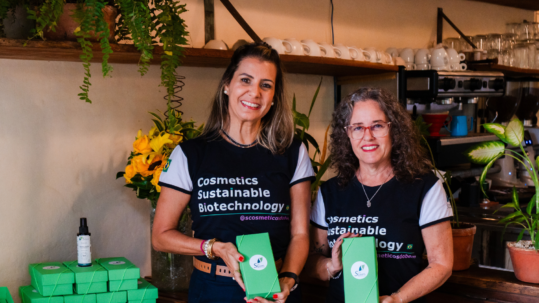 Foto colorida apresenta duas mulheres sorridentes, vestindo camisetas pretas com a frase "Cosmetics Sustainable Biotechnology", elas posam atrás de um balcão de madeira em um café, segurando caixas verdes de produtos. Ao redor delas, há embalagens empilhadas, flores amarelas e prateleiras com louças, criando um ambiente acolhedor e profissional. Fim da descrição.