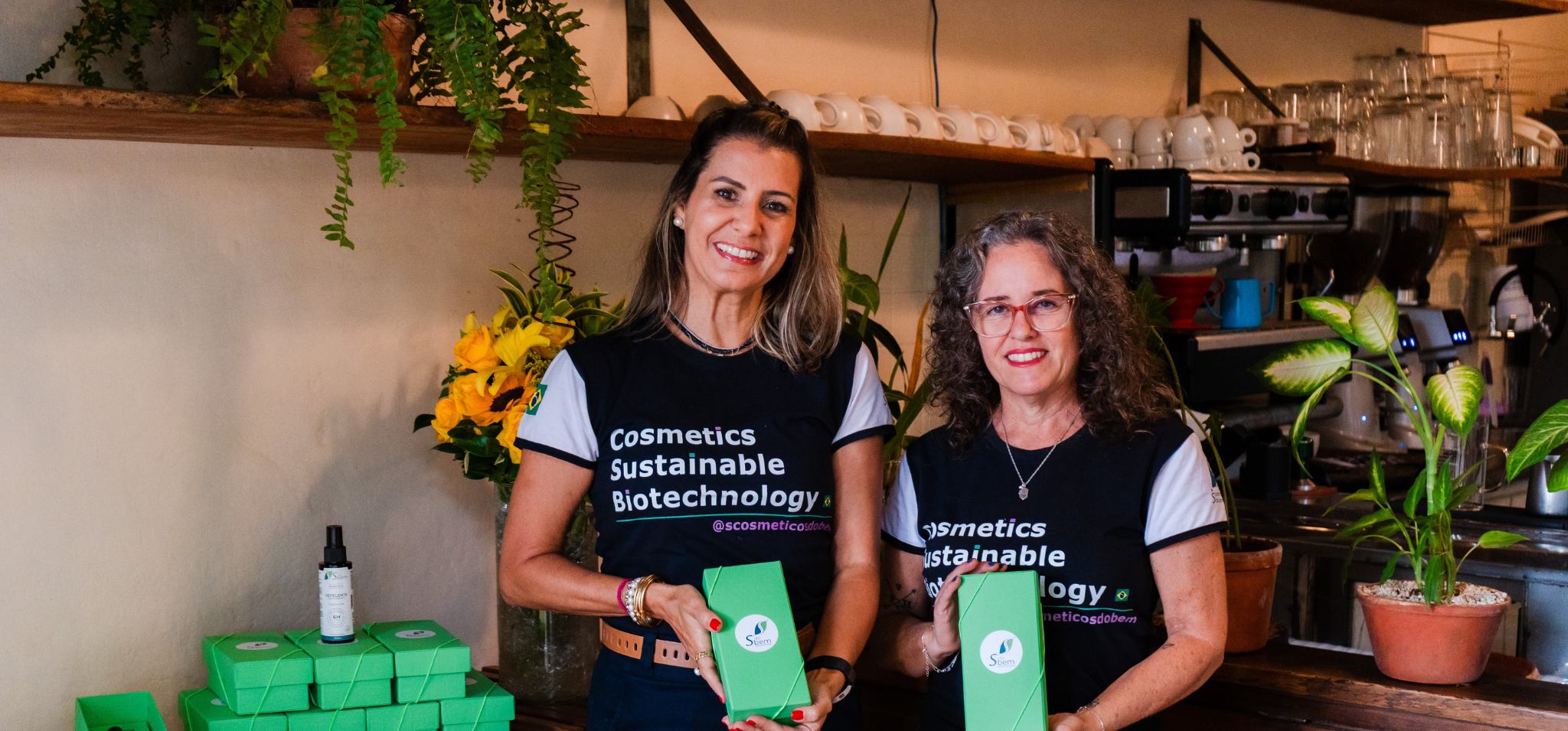Foto colorida apresenta duas mulheres sorridentes, vestindo camisetas pretas com a frase "Cosmetics Sustainable Biotechnology", elas posam atrás de um balcão de madeira em um café, segurando caixas verdes de produtos. Ao redor delas, há embalagens empilhadas, flores amarelas e prateleiras com louças, criando um ambiente acolhedor e profissional. Fim da descrição.