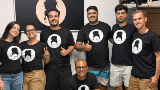 Foto colorida apresenta cinco homens e duas mulheres posando sorridentes para uma foto lado a lado. Todos vestem uma camisa preta contendo o logo da empresa-filha da Unicamp, Tieta.ai, que também aparece ao fundo da imagem. Fim da descrição.