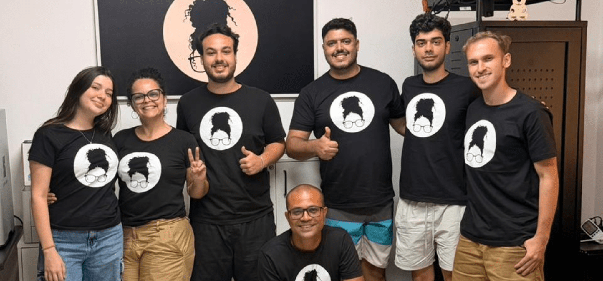 Foto colorida apresenta cinco homens e duas mulheres posando sorridentes para uma foto lado a lado. Todos vestem uma camisa preta contendo o logo da empresa-filha da Unicamp, Tieta.ai, que também aparece ao fundo da imagem. Fim da descrição.
