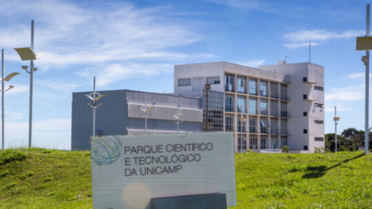 Foto colorida apresenta fachada do Parque Científico e Tecnológico da Unicamp, em Campinas, sob céu azul com nuvens esparsas. Em primeiro plano, sobre um gramado verde, há uma placa branca com o nome e o logotipo da instituição. Ao fundo, destaca-se um edifício moderno de dois blocos — um cinza mais baixo e outro branco de quatro andares com grandes janelas de vidro —, cercado por postes de iluminação contemporâneos. Fim da descrição.