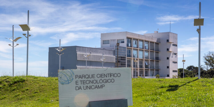 Foto colorida apresenta fachada do Parque Científico e Tecnológico da Unicamp, em Campinas, sob céu azul com nuvens esparsas. Em primeiro plano, sobre um gramado verde, há uma placa branca com o nome e o logotipo da instituição. Ao fundo, destaca-se um edifício moderno de dois blocos — um cinza mais baixo e outro branco de quatro andares com grandes janelas de vidro —, cercado por postes de iluminação contemporâneos. Fim da descrição.