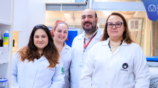 Fotografia colorida em plano médio de quatro pesquisadores — três mulheres e um homem sorrindo em um laboratório, todos vestindo jalecos brancos da Unicamp. Da esquerda para a direita, a primeira mulher tem cabelos castanhos longos e apoia a mão sobre uma bancada com equipamentos. Fim da descrição.