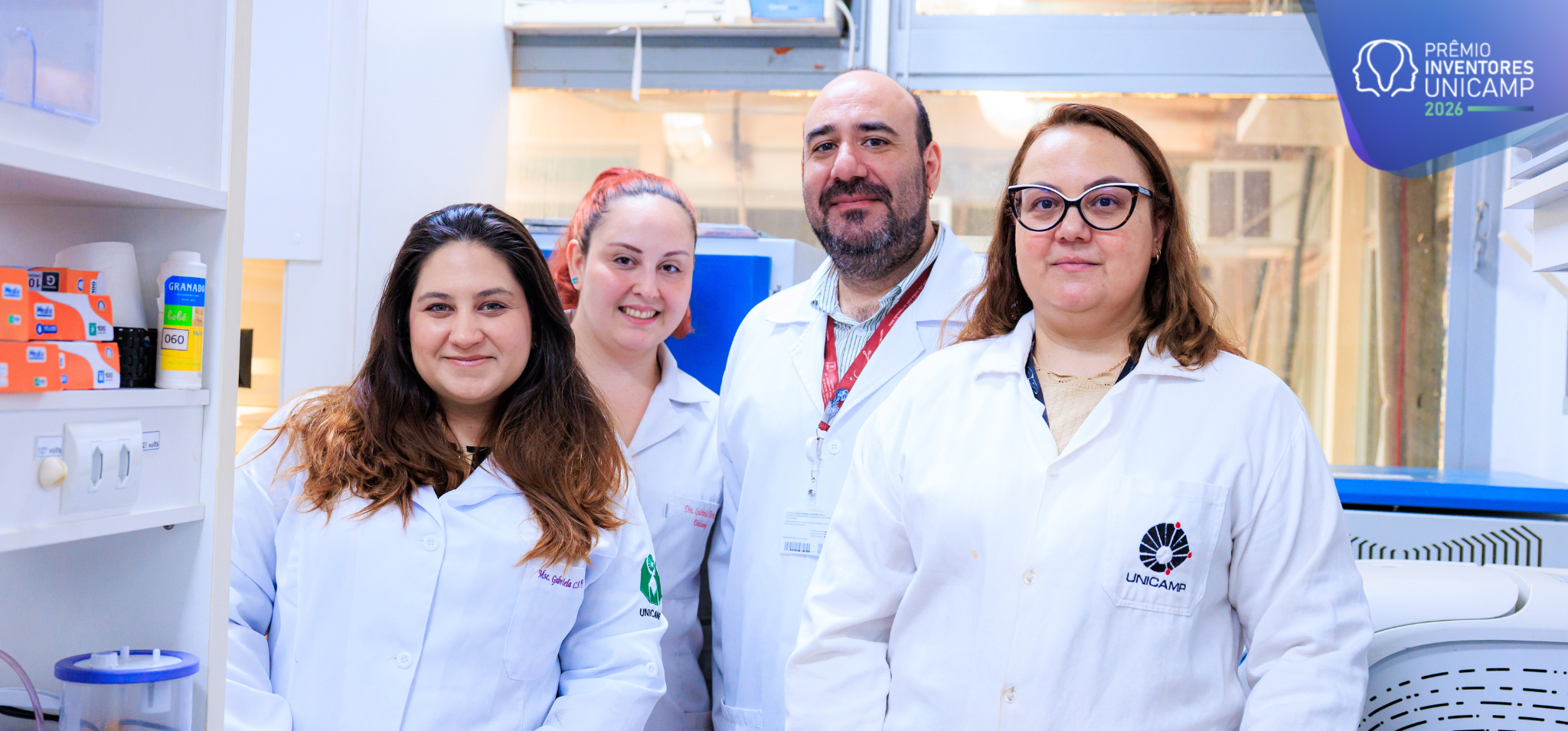 Fotografia colorida em plano médio de quatro pesquisadores — três mulheres e um homem sorrindo em um laboratório, todos vestindo jalecos brancos da Unicamp. Da esquerda para a direita, a primeira mulher tem cabelos castanhos longos e apoia a mão sobre uma bancada com equipamentos. Fim da descrição.
