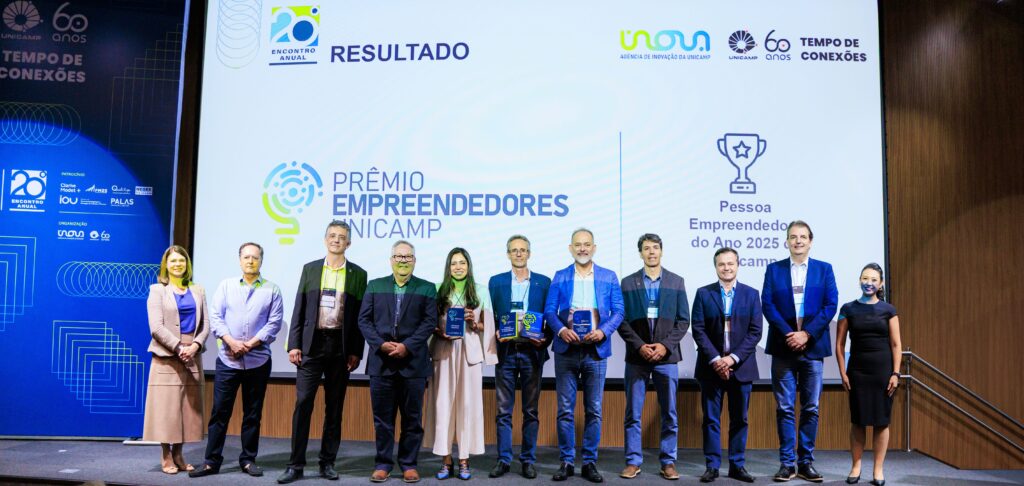 Uma fotografia de um palco de madeira onde 10 pessoas, incluindo sete homens e três mulheres, estão alinhadas lado a lado em uma cerimônia de premiação da Unicamp. Quatro pessoas seguram placas de acrílico translúcidas e coloridas com logotipos. Atrás delas, um grande telão de LED exibe o título principal "PRÊMIO EMPREENDEDORES UNICAMP" e o logotipo da premiação no centro. À direita do telão, sob um ícone de troféu, lê-se "Pessoa Empreendedora do Ano 2025". Logotipos da "Inova Unicamp" e da celebração "Unicamp 60 anos, Tempo de Conexões" são visíveis no topo do telão. À esquerda do palco, um painel vertical lista patrocinadores e exibe mais logos da Unicamp. Fim da descrição.
