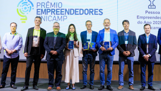 Um grupo de dez pessoas está em pé num palco diante de uma tela de projeção onde se lê "10º Prêmio Empreendedores Unicamp". A pessoa mais à esquerda é uma mulher com um blazer bege longo, uma blusa azul e uma saia longa bege. Ao lado dela está um homem com uma camisa de botões azul-clara listrada e calças pretas. A próxima pessoa é um homem com um paletó preto e calças pretas. A próxima pessoa é um homem com um paletó marrom-escuro e calças cinza-escuras. A próxima pessoa é uma mulher com um paletó creme e calças largas creme. Ela está segurando um troféu retangular azul e prateado. A próxima pessoa é um homem com um paletó azul-marinho e jeans azuis. Ele está segurando um troféu verde. A próxima pessoa é um homem com um paletó azul-médio e jeans azuis. Ele está segurando um troféu azul e prateado. A próxima pessoa é um homem com um paletó marrom-escuro e jeans azuis. A próxima pessoa é um homem com um paletó preto e jeans pretos. A pessoa mais à direita é um homem com um paletó azul e calças cinza. O palco tem painéis de madeira texturizada ao longo da base e da lateral. fim da descrição