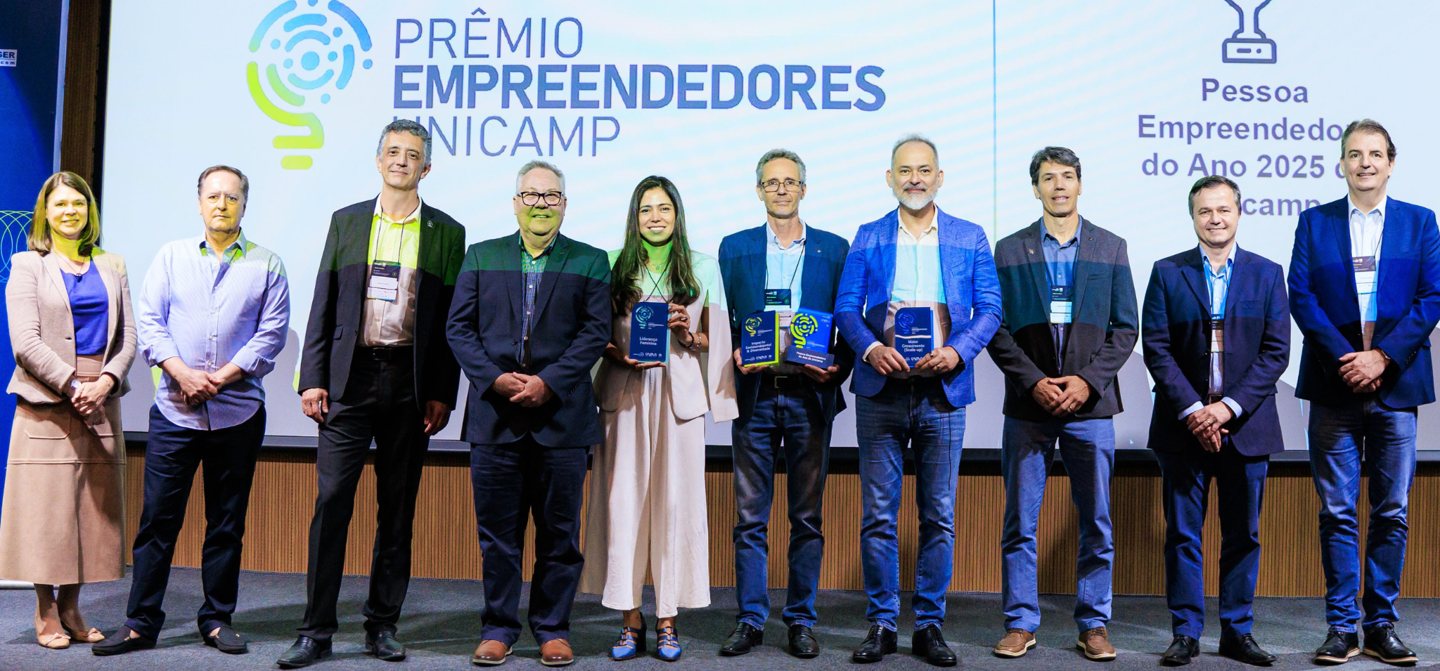 Um grupo de dez pessoas está em pé num palco diante de uma tela de projeção onde se lê "10º Prêmio Empreendedores Unicamp". A pessoa mais à esquerda é uma mulher com um blazer bege longo, uma blusa azul e uma saia longa bege. Ao lado dela está um homem com uma camisa de botões azul-clara listrada e calças pretas. A próxima pessoa é um homem com um paletó preto e calças pretas. A próxima pessoa é um homem com um paletó marrom-escuro e calças cinza-escuras. A próxima pessoa é uma mulher com um paletó creme e calças largas creme. Ela está segurando um troféu retangular azul e prateado. A próxima pessoa é um homem com um paletó azul-marinho e jeans azuis. Ele está segurando um troféu verde. A próxima pessoa é um homem com um paletó azul-médio e jeans azuis. Ele está segurando um troféu azul e prateado. A próxima pessoa é um homem com um paletó marrom-escuro e jeans azuis. A próxima pessoa é um homem com um paletó preto e jeans pretos. A pessoa mais à direita é um homem com um paletó azul e calças cinza. O palco tem painéis de madeira texturizada ao longo da base e da lateral. fim da descrição