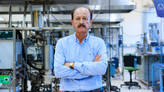 Uma fotografia colorida na horizontal em plano médio, do quadril para cima. O homem está no centro do ambiente de um laboratório com os braços cruzados, de pé. Fim da descrição.