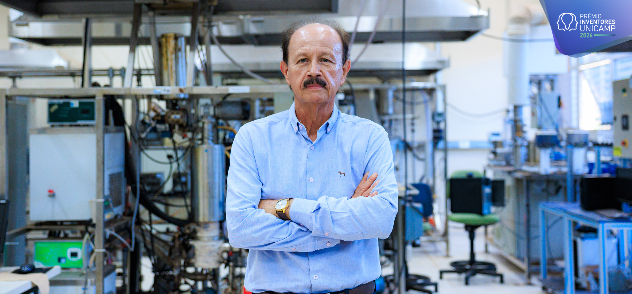 Uma fotografia colorida na horizontal em plano médio, do quadril para cima. O homem está no centro do ambiente de um laboratório com os braços cruzados, de pé. Fim da descrição.