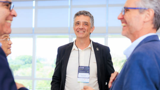 Fotografia de um grupo de homens conversando e sorrindo durante o 21º Encontro Anual Unicamp Ventures. No centro da imagem e voltado para a frente, há um homem de cabelos grisalhos curtos, vestindo uma camisa clara e um blazer preto. Ele usa um crachá do evento no pescoço onde se lê o nome "Renato Lopes" e sorri de forma atenta. Em primeiro plano, à esquerda e à direita, aparecem de perfil outros dois homens que participam da roda de conversa; ambos usam óculos, vestem camisas e paletós em tons de azul e também estão sorrindo. O ambiente é bem iluminado, com grandes janelas de vidro ao fundo que mostram a luz do dia e o reflexo de árvores desfocadas. Fim da descrição.
