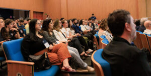Fotografia de um auditório com várias pessoas sentadas durante o 21º Encontro Anual Unicamp Ventures. O público, composto por homens e mulheres, está sentado em fileiras de poltronas azuis com encostos de madeira, olhando atentamente em direção ao lado esquerdo da imagem, onde possivelmente acontece uma apresentação. O ambiente possui paredes altas e teto revestidos com painéis de madeira clara. Em primeiro plano, à esquerda, destaca-se uma jovem de óculos usando blusa preta e calça cor de ferrugem, com uma bolsa preta apoiada ao seu lado. À direita, também em primeiro plano mas fora de foco, aparece a silhueta das costas e do perfil de um homem vestindo camisa preta. Fim da descrição.