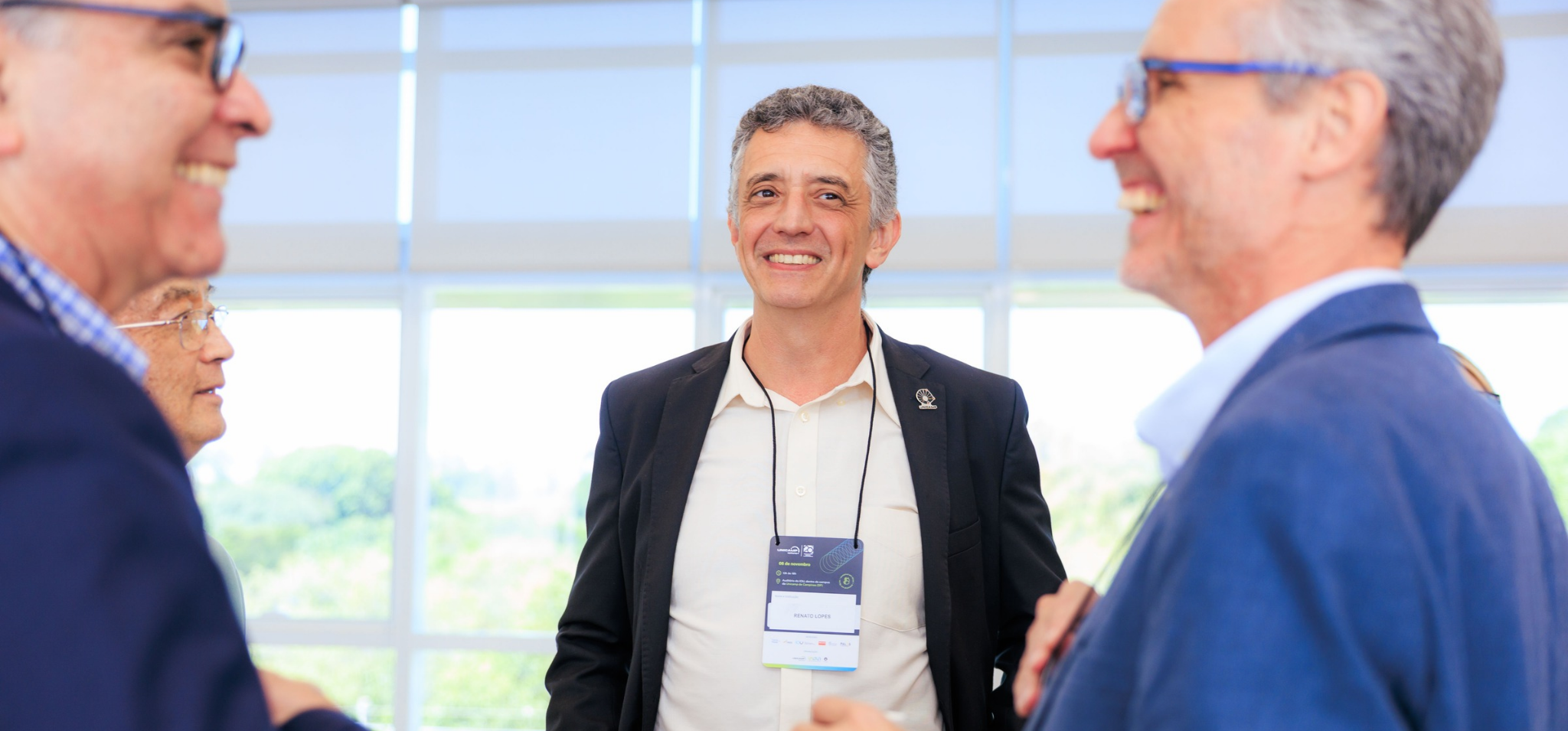 Fotografia de um grupo de homens conversando e sorrindo durante o 21º Encontro Anual Unicamp Ventures. No centro da imagem e voltado para a frente, há um homem de cabelos grisalhos curtos, vestindo uma camisa clara e um blazer preto. Ele usa um crachá do evento no pescoço onde se lê o nome "Renato Lopes" e sorri de forma atenta. Em primeiro plano, à esquerda e à direita, aparecem de perfil outros dois homens que participam da roda de conversa; ambos usam óculos, vestem camisas e paletós em tons de azul e também estão sorrindo. O ambiente é bem iluminado, com grandes janelas de vidro ao fundo que mostram a luz do dia e o reflexo de árvores desfocadas. Fim da descrição.