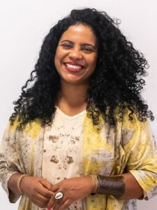 A imagem apresenta uma mulher de pele escura e cabelos escuros encaracolados a sorrir amplamente para a câmara, usando batom vermelho. Veste uma blusa clara sob um casaco largo com padrões em tons de amarelo, cinzento e branco. Destacam-se os seus acessórios: uma bracelete metálica larga no pulso esquerdo, pulseiras de fio no pulso direito e um grande anel com formato de búzio. O fundo é uma parede branca lisa. Fim da descrição. 
