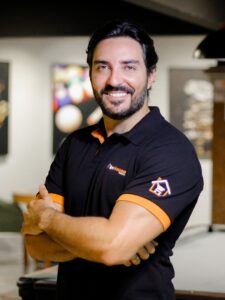 A imagem mostra um homem de cabelo escuro, barba e bigode a sorrir para a câmara, de braços cruzados. Ele veste um polo preto com detalhes em cor de laranja na gola e nas bainhas das mangas, ostentando o logótipo "InHouse MARKET" no peito e um símbolo com uma casa e um carrinho de compras na manga esquerda. O fundo está desfocado, aparentando ser um espaço de convívio interior com uma mesa de bilhar. Fim da descrição. 