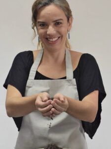 A fotografia exibe uma mulher de pele clara e cabelo apanhado a sorrir para a câmara. Ela veste uma camisola preta e um avental cinzento claro, posando com as mãos em forma de concha, de onde deixa cair pequenos grãos ou sementes escuras. O fundo da imagem é liso e numa tonalidade muito clara. Fim da descrição. 