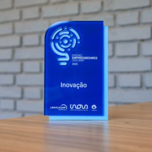 A imagem mostra um troféu de acrílico azul sobre uma mesa de madeira, com uma parede de tijolo branco ao fundo. O troféu tem um logótipo vazado na parte superior, as inscrições "PRÊMIO EMPREENDEDORES UNICAMP 2025" e a categoria "Inovação", apoiando-se numa placa traseira azul clara. Na base, estão os logótipos da UNICAMP ventures, Inova Unicamp e Unicamp. Fim da descrição. 