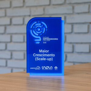 A imagem exibe um troféu de acrílico azul pousado numa mesa de madeira, tendo como fundo uma parede de tijolo branco. O prémio apresenta um logótipo vazado, as palavras "PRÊMIO EMPREENDEDORES UNICAMP 2025", a categoria "Maior Crescimento (Scale-up)" e uma placa traseira de cor azul clara. A base contém os logótipos da UNICAMP ventures, Inova Unicamp e Unicamp. Fim da descrição. 