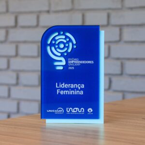 A imagem apresenta um troféu de acrílico azul numa mesa de madeira, contra uma parede de tijolo branco. Este troféu destaca a categoria "Liderança Feminina", juntamente com a inscrição "PRÊMIO EMPREENDEDORES UNICAMP 2025" e um logótipo circular vazado, tendo por trás uma placa azul clara. Na base encontram-se os logótipos da UNICAMP ventures, Inova Unicamp e Unicamp. Fim da descrição. 
