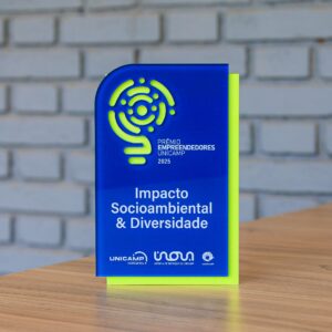 A imagem mostra um troféu de acrílico azul sobre uma mesa de madeira, com uma parede de tijolo branco em segundo plano. O troféu exibe a categoria "Impacto Socioambiental & Diversidade", a inscrição "PRÊMIO EMPREENDEDORES UNICAMP 2025" e um logótipo vazado que revela uma placa traseira de cor verde néon. Os logótipos da UNICAMP ventures, Inova Unicamp e Unicamp figuram na base. Fim da descrição. 